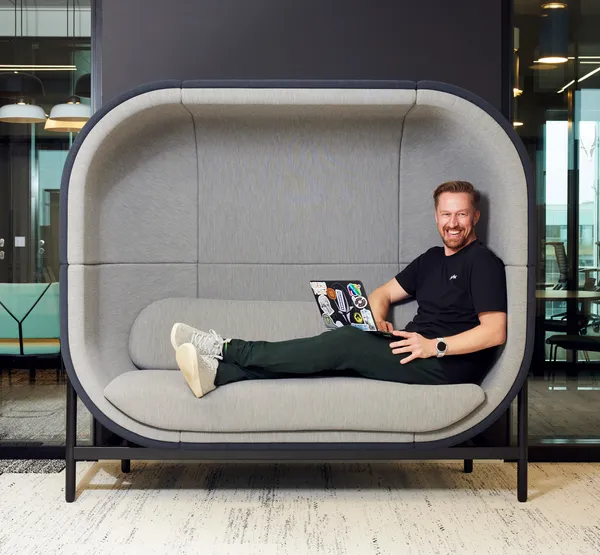 Guido Jansen, a CRO and UX specialist, sitting casually on a modern grey office couch with a laptop. He's wearing casual business attire and has a welcoming expression, representing his approachable consulting style.